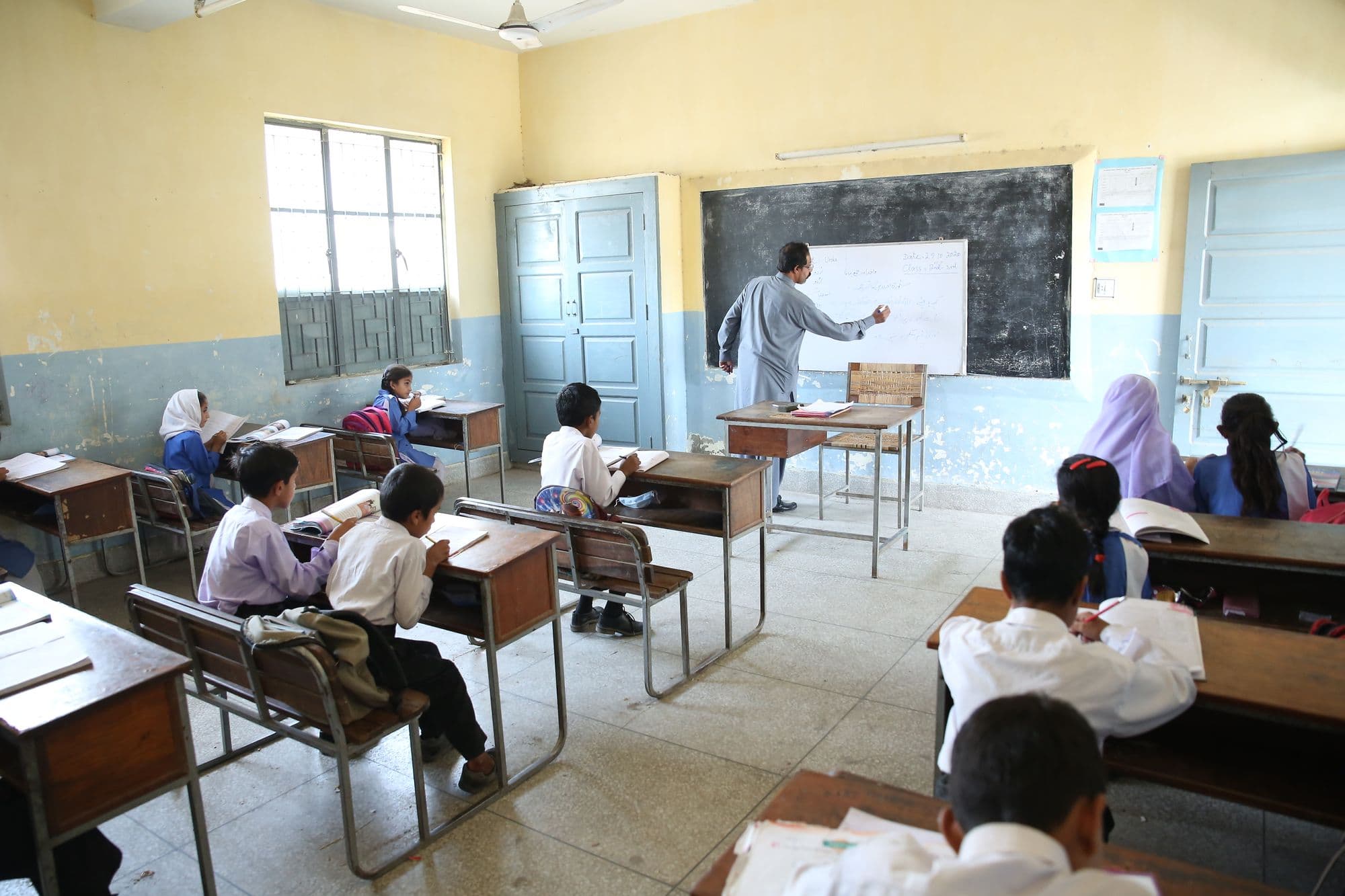 A teacher writing on a whiteboard at the front of a classroom while students work at their desks