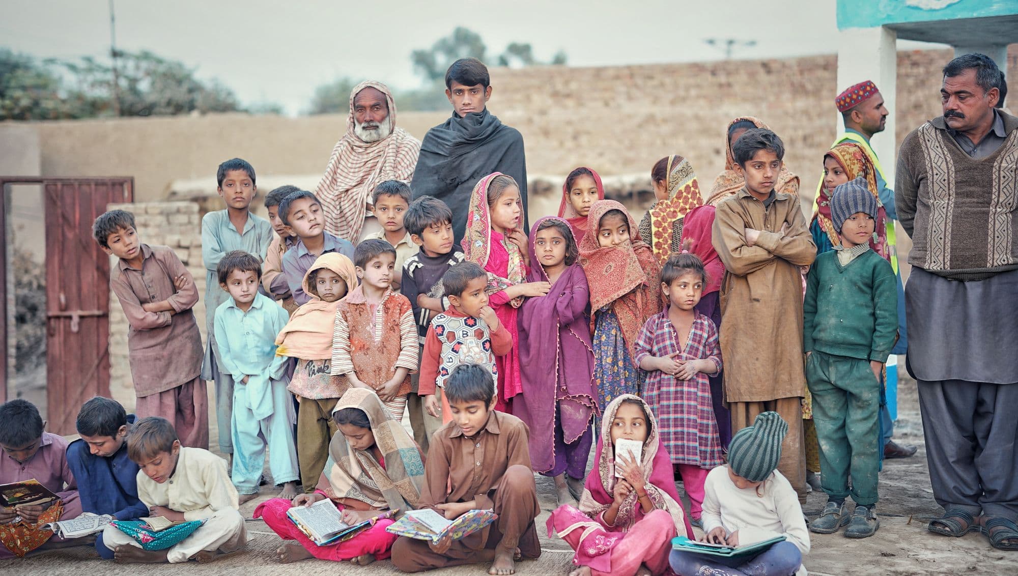 Children and adults gathered outdoors in a village, some of the children sitting on the ground reading books
