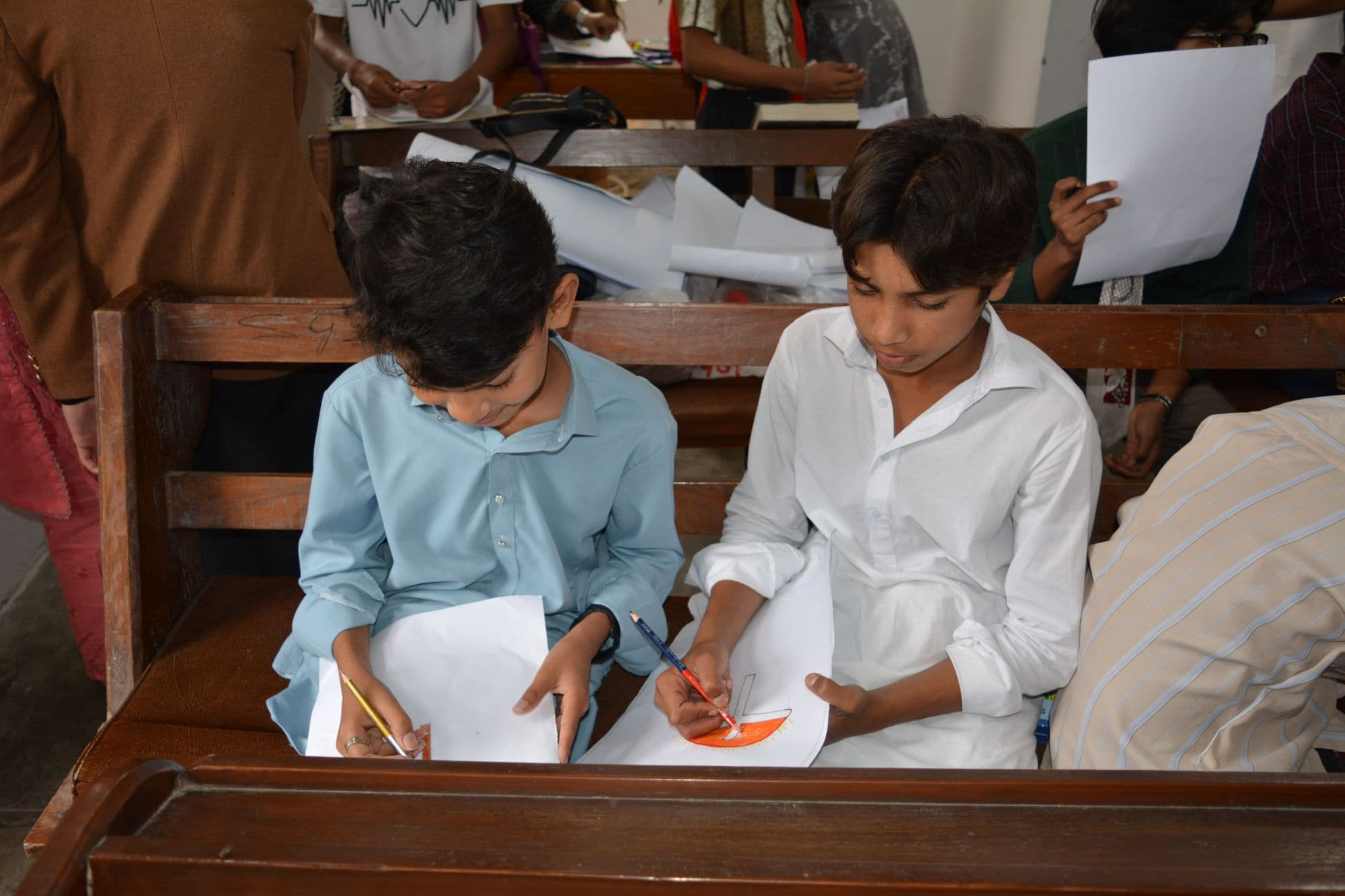 Two students sitting at a wooden desk, drawing and writing on sheets of paper